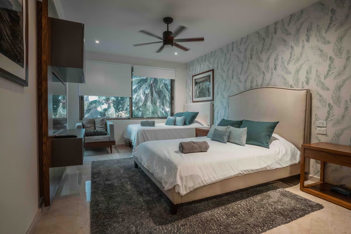 This peaceful guest room at Villa Patrón Cancun offers two plush double beds, soft tropical tones, and a view framed by lush palm trees — perfect for friends or family. | Esta acogedora habitación de Villa Patrón Cancún ofrece dos camas matrimoniales, tonos tropicales suaves y una vista enmarcada por palmeras, ideal para amigos o familia.
