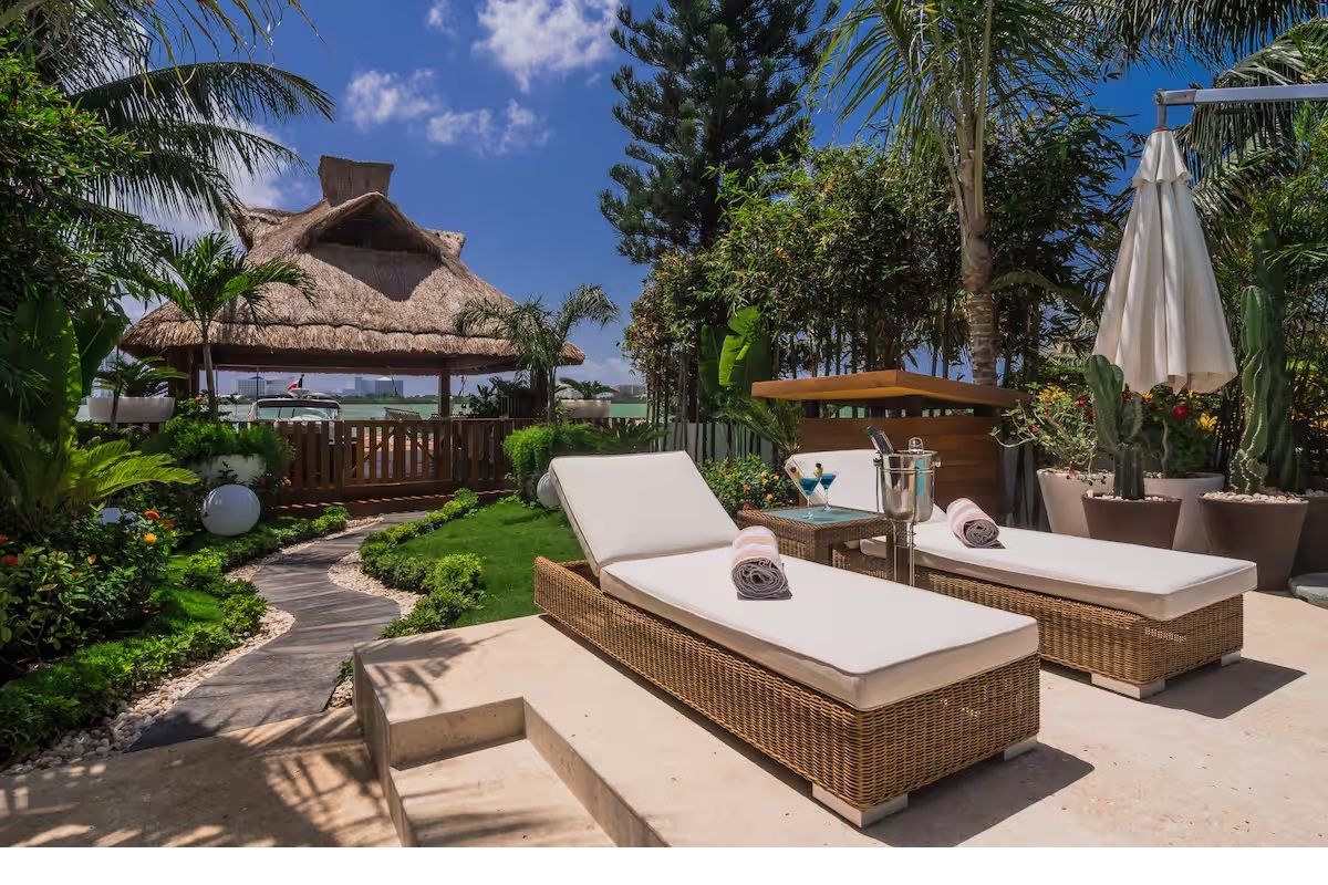 Relax by the lagoon under the palapa at Villa Patrón, surrounded by lush gardens, sun loungers, and serene Caribbean views. | Relájate frente a la laguna bajo la palapa de Villa Patrón, rodeado de jardines tropicales, camastros y vistas al Caribe.
