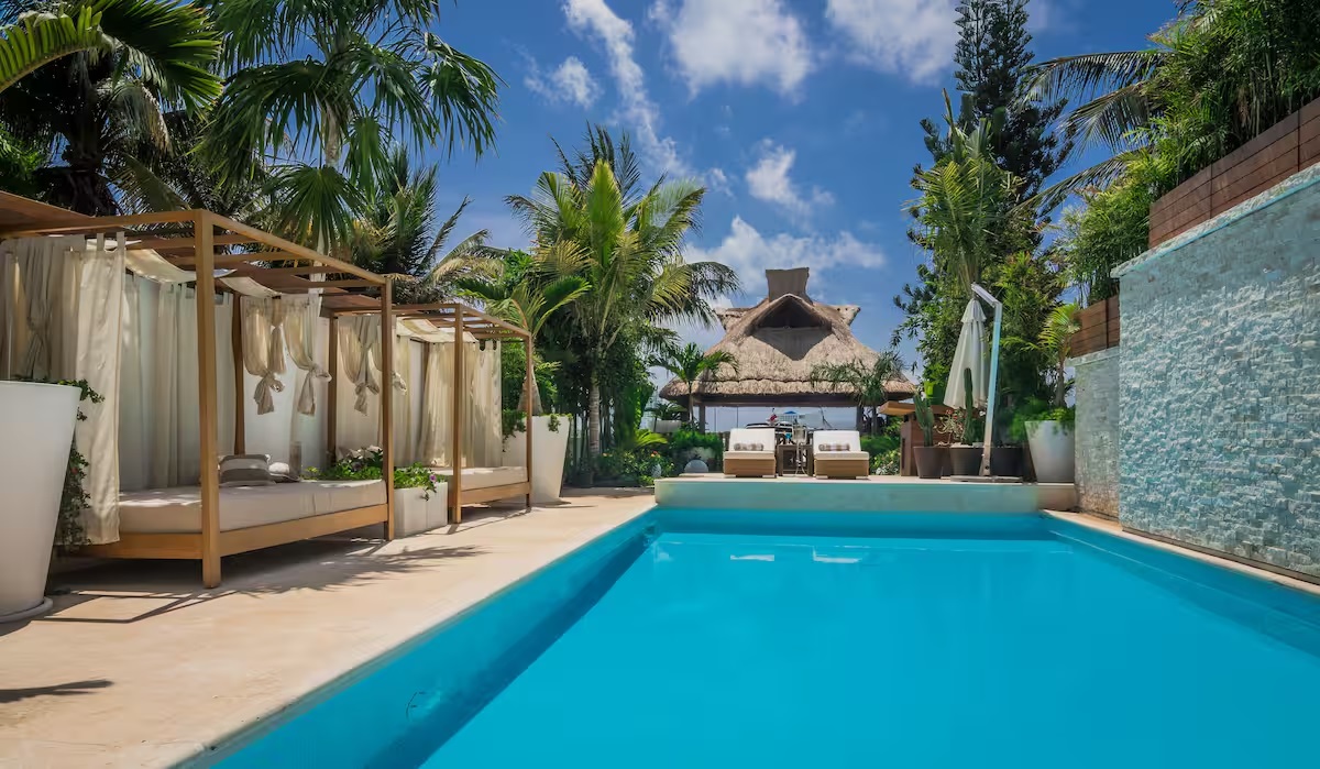 Villa Patrón’s main pool framed by tropical palms, elegant cabanas, and a thatched palapa overlooking Nichupté Lagoon — luxury and serenity in perfect harmony. | La alberca principal de Villa Patrón rodeada de palmeras, camas balinesas y una palapa con vista a la Laguna Nichupté — lujo y serenidad en perfecta armonía.
