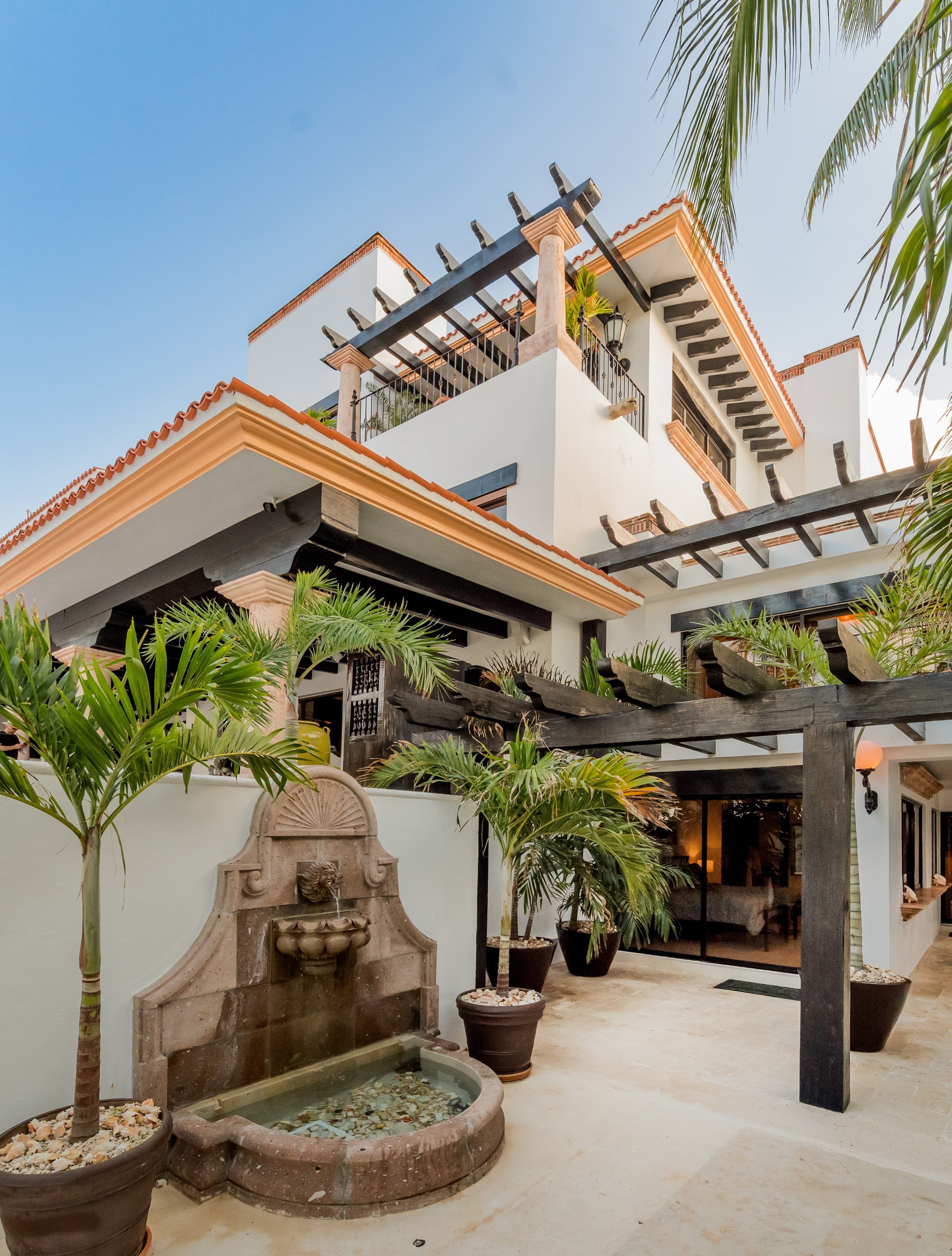 Elegant villa exterior with tropical palms and stone fountain — Villa Albatros, part of I Live Where You Vacation’s Riviera Maya luxury collection. | Exterior elegante con palmeras tropicales y fuente de piedra — Villa Albatros, parte de la colección de lujo Riviera Maya de I Live Where You Vacation.
