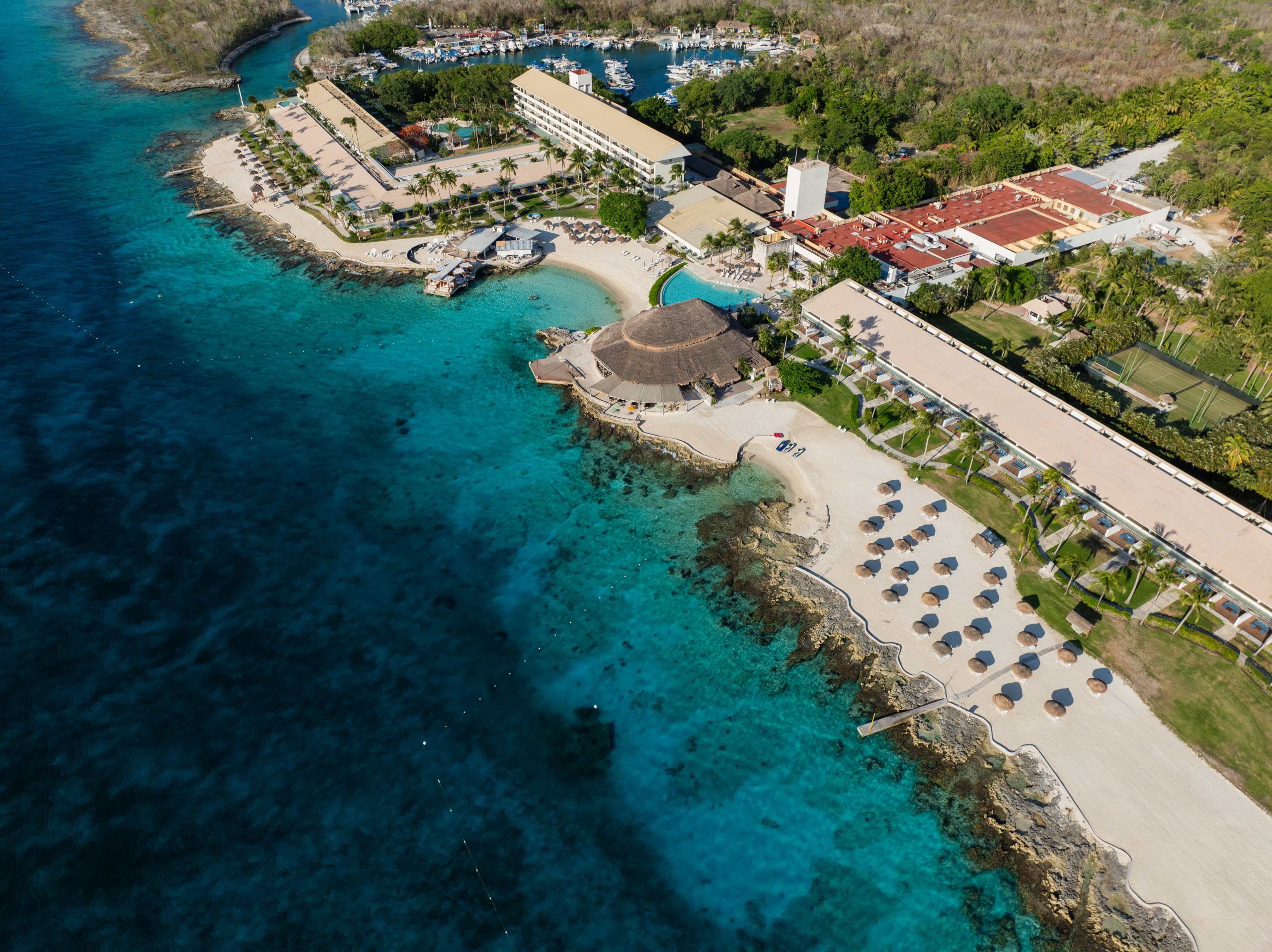 Aerial view of Cozumel Island resort and turquoise Caribbean waters, featuring beach umbrellas and coral reefs by I Live Where You Vacation. | Vista aérea de un resort en Isla Cozumel con aguas turquesas del Caribe, sombrillas de playa y arrecifes de coral por I Live Where You Vacation.
