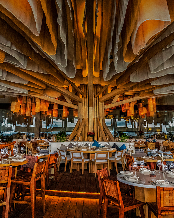 Interior view of Ilios Cancun, a fine dining restaurant in the Riviera Maya, showcasing an impressive wooden ceiling structure resembling a tree with warm ambient lighting and elegantly set tables — a masterpiece of contemporary design and luxury dining. – I Live Where You Vacation | Vista interior de Ilios Cancún, un restaurante de alta cocina en la Riviera Maya, con una impresionante estructura de techo de madera que evoca un árbol, iluminación cálida y mesas elegantemente dispuestas — una obra maestra de diseño contemporáneo y gastronomía de lujo. – I Live Where You Vacation