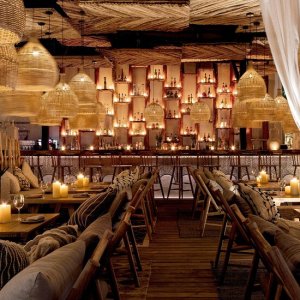 Warmly lit interior of Chambao Cancun, showcasing a luxurious wooden dining space with woven pendant lights, flickering candles, and an illuminated bar lined with fine bottles — an atmosphere of refined coastal elegance – I Live Where You Vacation | Interior cálidamente iluminado de Chambao Cancún, que muestra un lujoso espacio de comedor de madera con lámparas colgantes tejidas, velas encendidas y un bar iluminado con botellas selectas — una atmósfera de elegancia costera refinada – I Live Where You Vacation